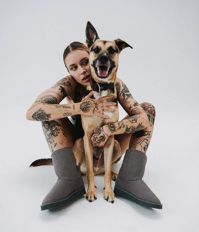 Woman with tattoos sitting on the floor with a dog, wearing gray vegan boots.
