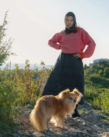 Woman in a pink top and navy blue skirt standing with a dog on a rocky path with greenery in the background