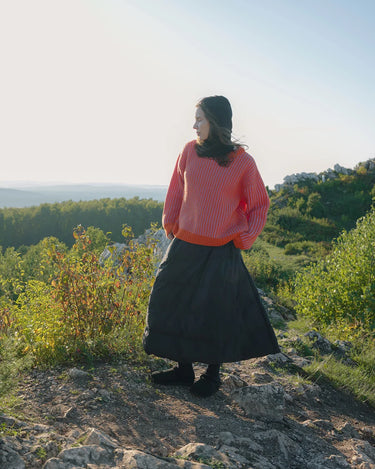 Person wearing a pink sweater and navy blue skirt standing on a rocky outcrop with greenery and a clear sky in the background.