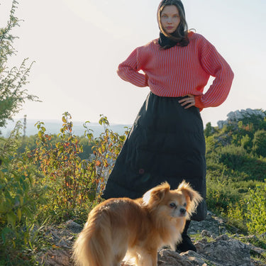 Woman in a pink top and navy blue skirt standing with a dog on a rocky path with greenery in the background